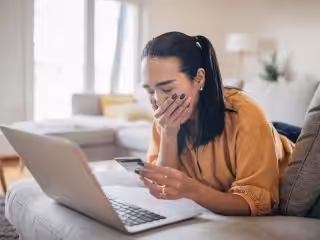 Eine junge Frau, die gestresst aussieht, während sie auf einem Sofa mit Laptop und Smartphone sitzt, im hellen Wohnzimmer mit natürlichen Lichtverhältnissen, möglicherweise Probleme mit Technik oder Streamingdiensten, modernes Home-Entertainment System, entspannte Wohnatmosphäre.