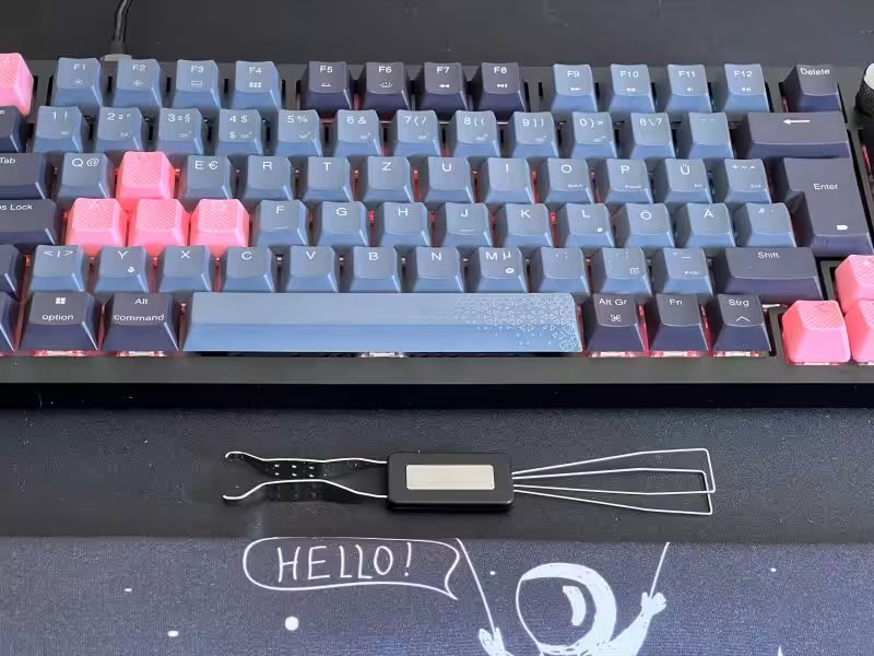 Leichter mechanischer Tastatur mit blauen und pinken Tasten, daneben ein digitales Messgerät mit Drähten, auf einem schwarzen Surface mit "HELLO!" Zeichnung.