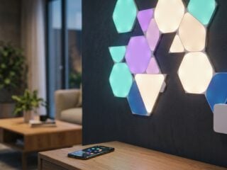 Create a sleek editorial product photograph of a modern smart home setup: a geometric wall installation of hexagonal and triangular illuminated panels mounted on a dark accent wall, glowing in soft ambient colors like teal, purple, and warm white. Focus on a small, minimalist white adapter module connected discreetly at the edge of the panel installation, with a subtle cable running to it. In the background, blurred, show a smartphone on a nearby table displaying a smart lighting app interface with simple colored dots representing zones. The lighting should feel warm and inviting, shot in a contemporary living room setting with soft shadows and a shallow depth of field. Style: clean tech-lifestyle photography, high production value, realistic textures, cinematic color grading with cool-to-warm contrast, professional studio quality, no visible text or logos anywhere in the image.