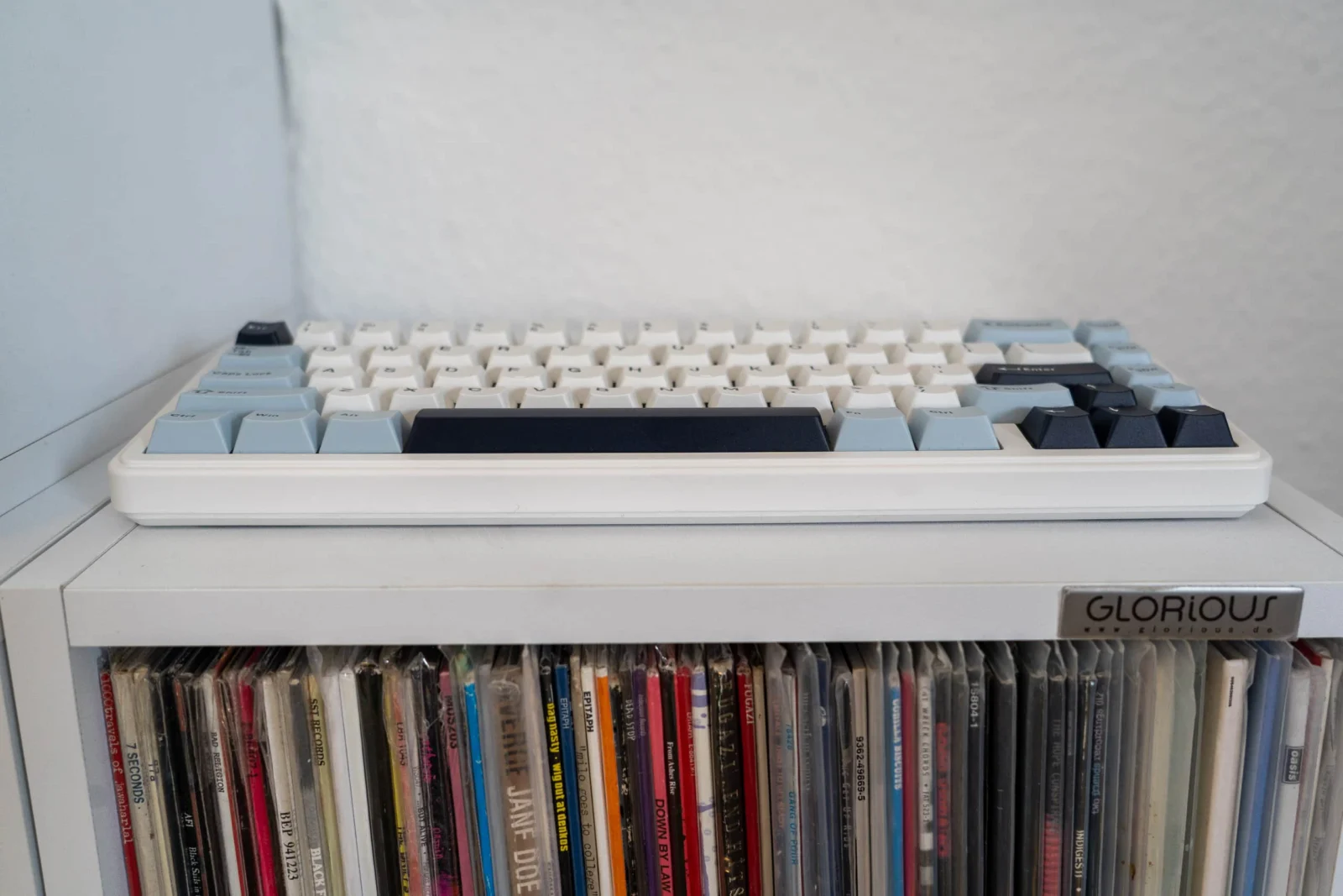 Günstig, gasket, geil? Was die EPOMAKER x AULA F65 wirklich kann Hintergrundbild mit mechanischer Tastatur mit blauen, weißen und schwarzen Tasten auf einem Bücherregal.