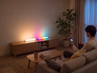 SwitchBot Lightbar: zwei Leisten liegen auf einem Lowboard im Wohnzimmer, ein Mann stellt die Farbe per App ein (Bild: SwitchBot)